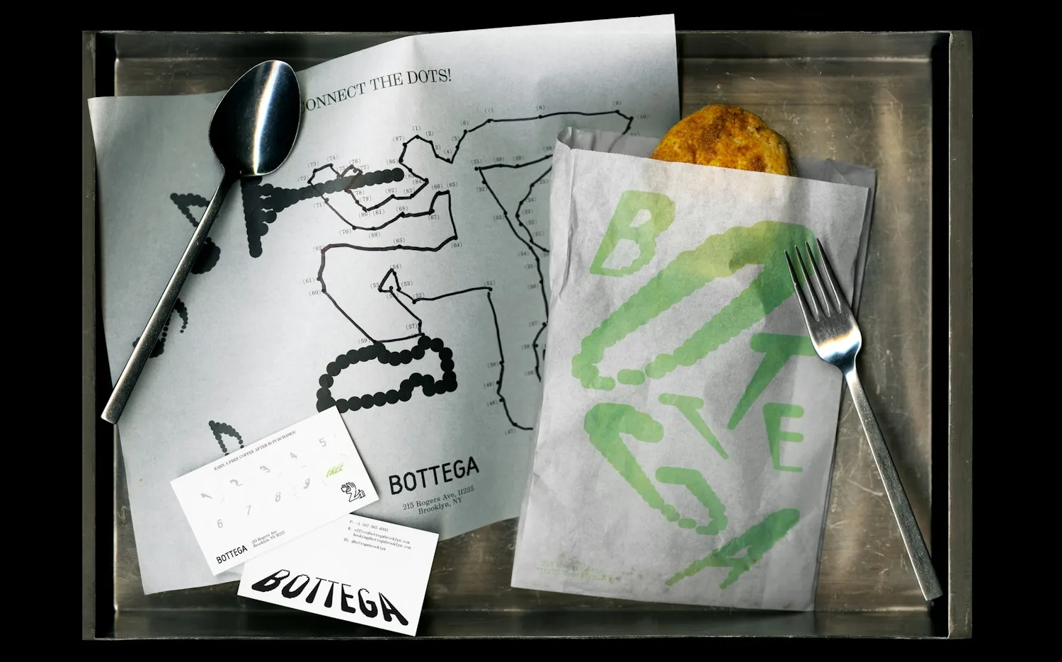 Bandeja metálica con papel de envolver, bolsa de papel y tarjetas de Bottega: las letras del nombre están formadas por cadenas de puntos verdes y negros que se hinchan y se deforman, junto a una cuchara, un tenedor y un pan.